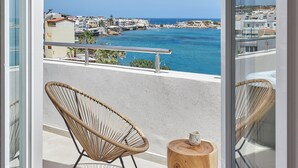 A balcony with a wicker chair and a wooden table, overlooking a coastal town with buildings and the sea. - Lotus Rise Hotel (Hersonissos)