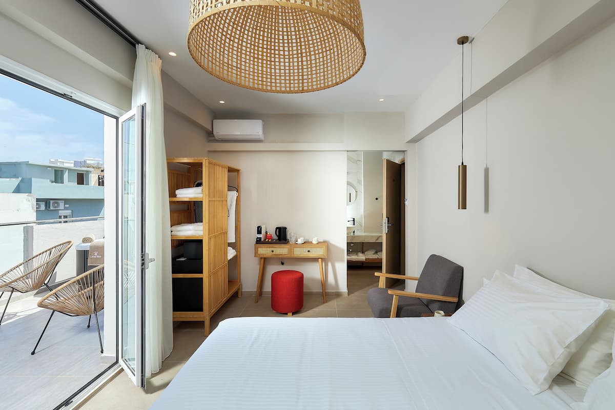 A modern hotel room with a large bed, a desk, a wardrobe, and a balcony with wicker chairs.
