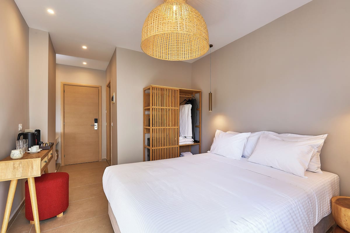 A hotel room with a large bed, a wooden wardrobe, a desk with a kettle, and a red stool.