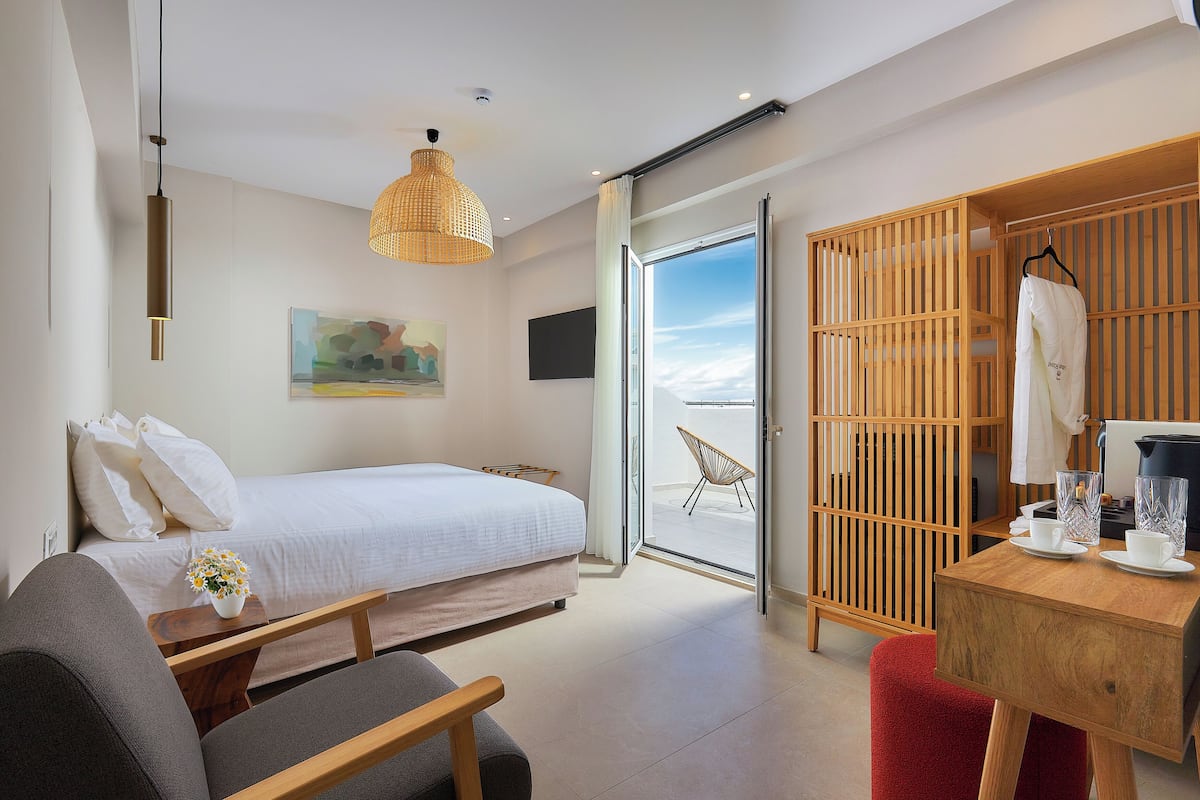 A modern hotel room with a bed, a desk, a chair, a hanging light fixture, and a sliding glass door leading to a balcony.