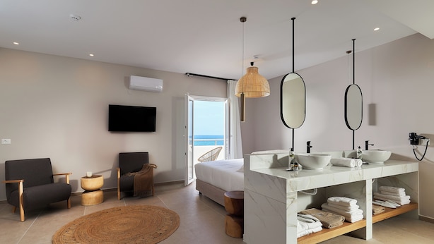 A modern bedroom with a bed, two chairs, a TV, a large window, and a bathroom with a sink and towel rack. - Lotus Rise Hotel (Hersonissos)
