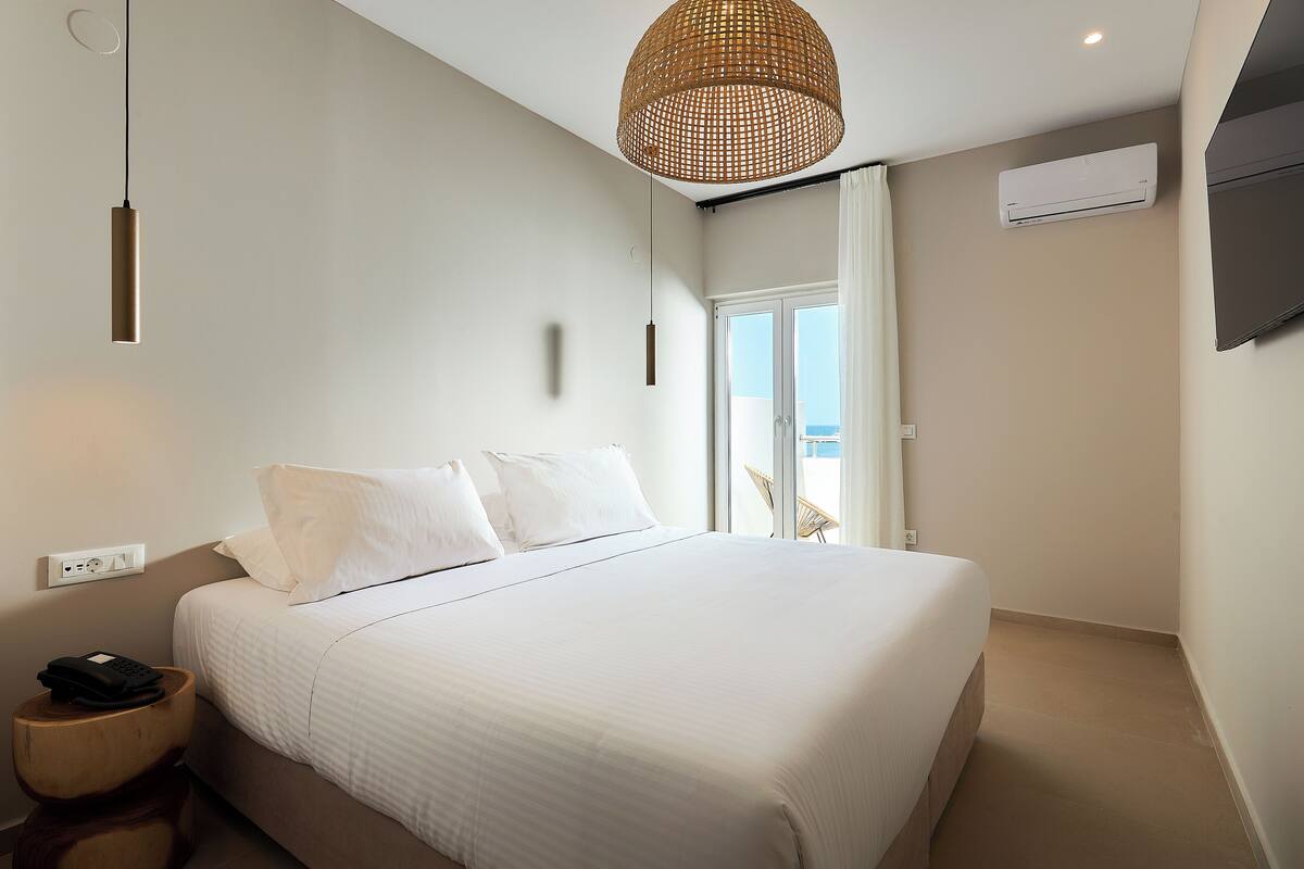 A hotel room with a bed, a hanging light fixture, an air conditioning unit, and a view of the outdoors.