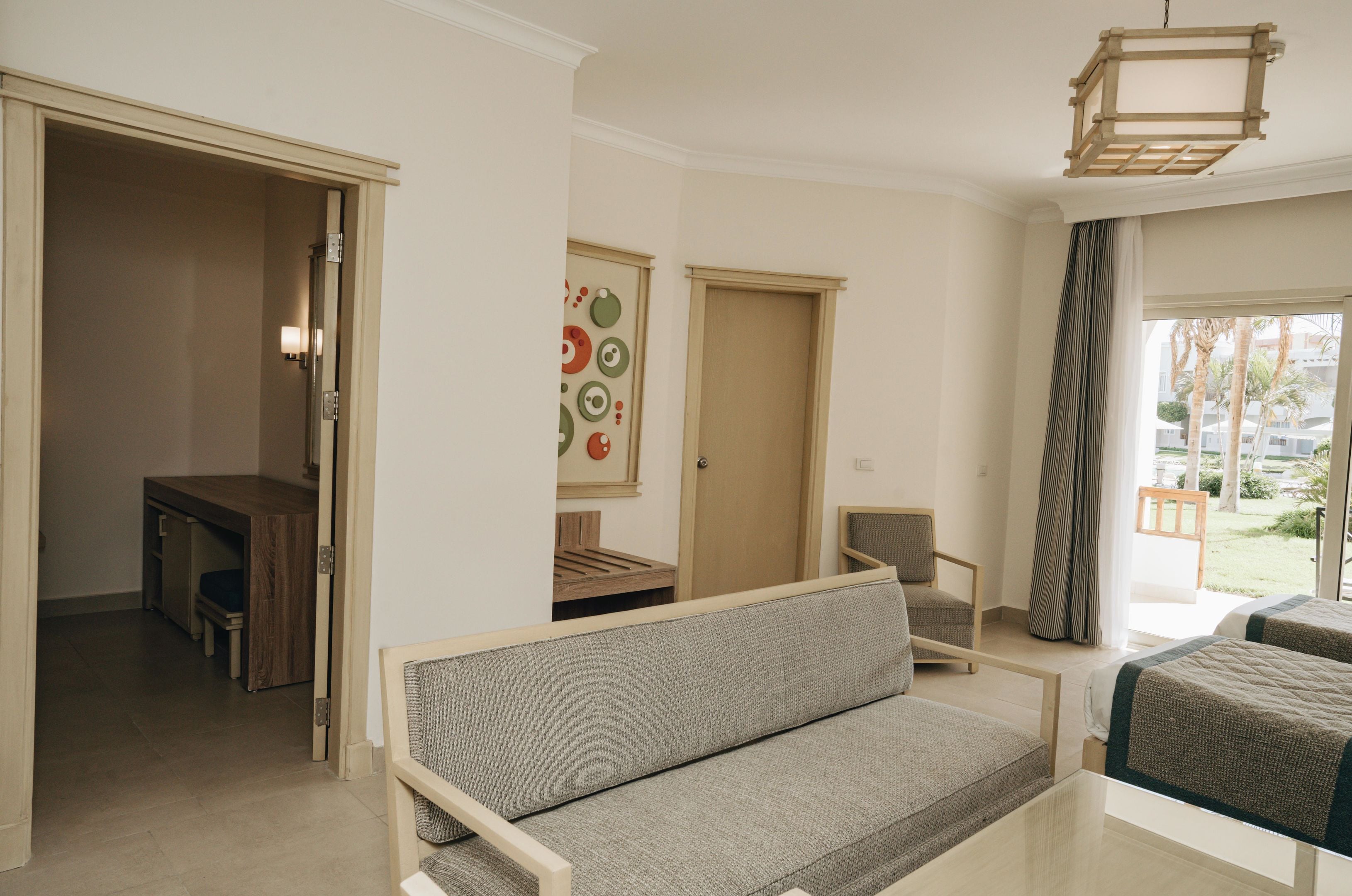 A room with a sofa, a desk, a chair, and a bed. There is a door leading to another room and a window with curtains.