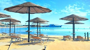 Uma praia com espreguiçadeiras e guarda-sóis de palha, um iate ao fundo, e um céu azul e límpido.