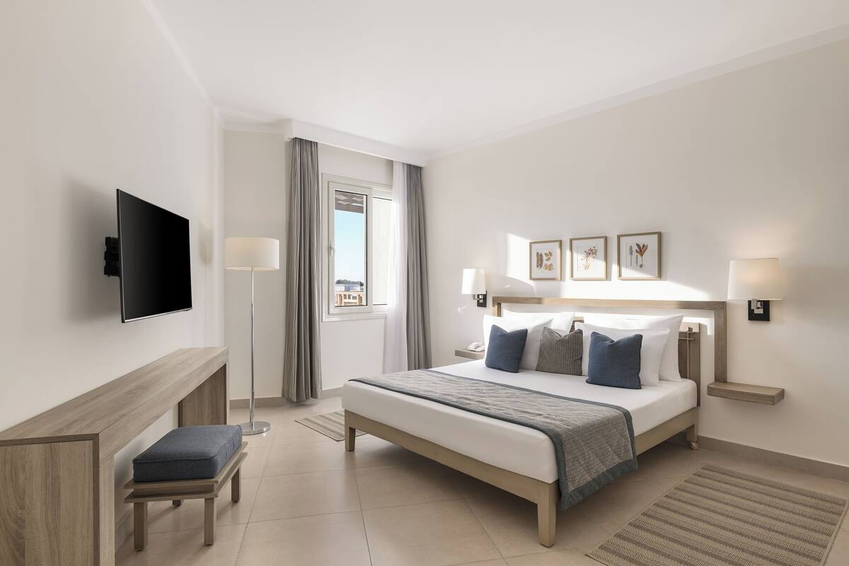 A modern hotel room with a bed, bedside tables, a flat-screen TV, a bench, and a window with curtains.