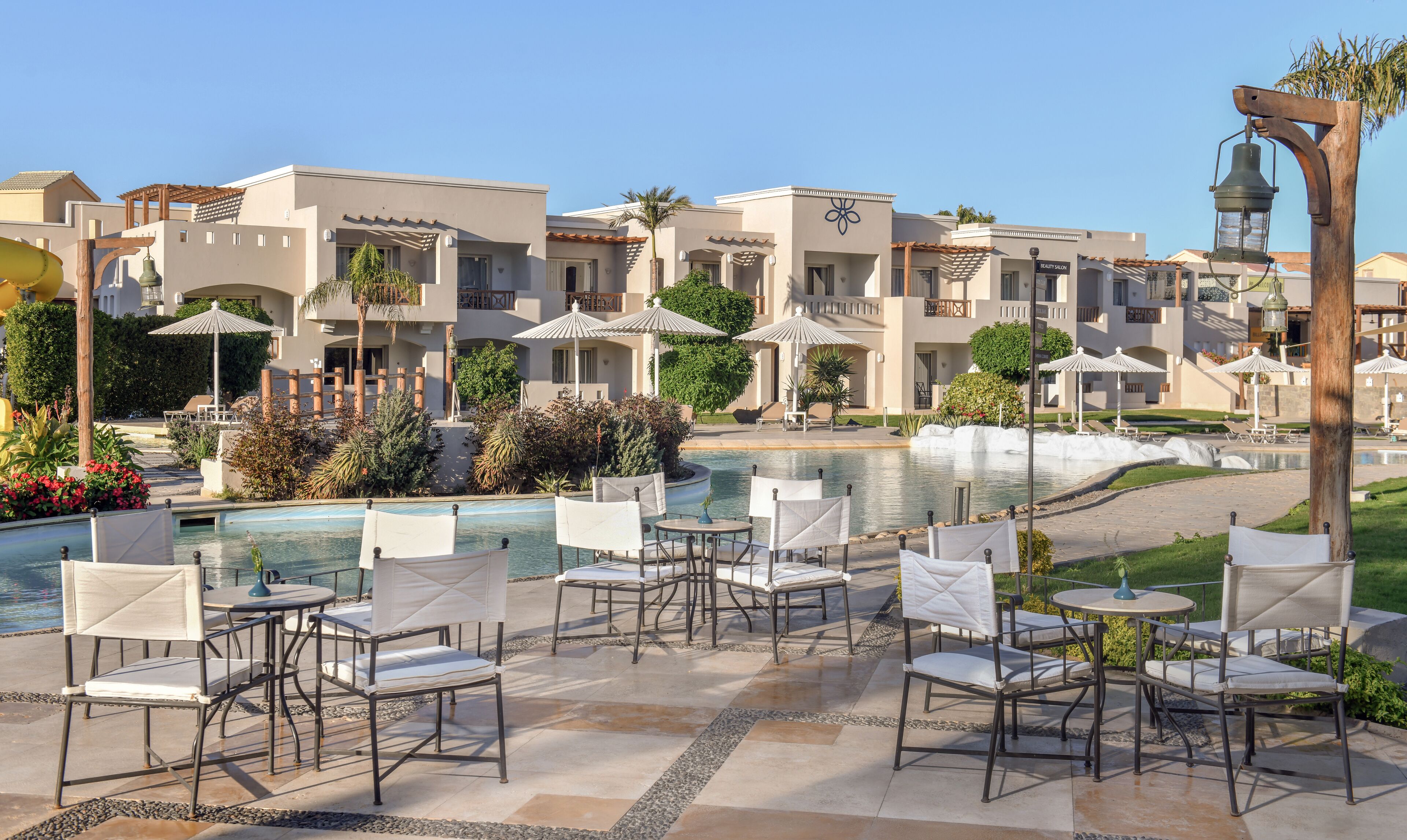 A poolside area with lounge chairs and umbrellas, a fountain, and a building with balconies and a sign for a beauty salon.