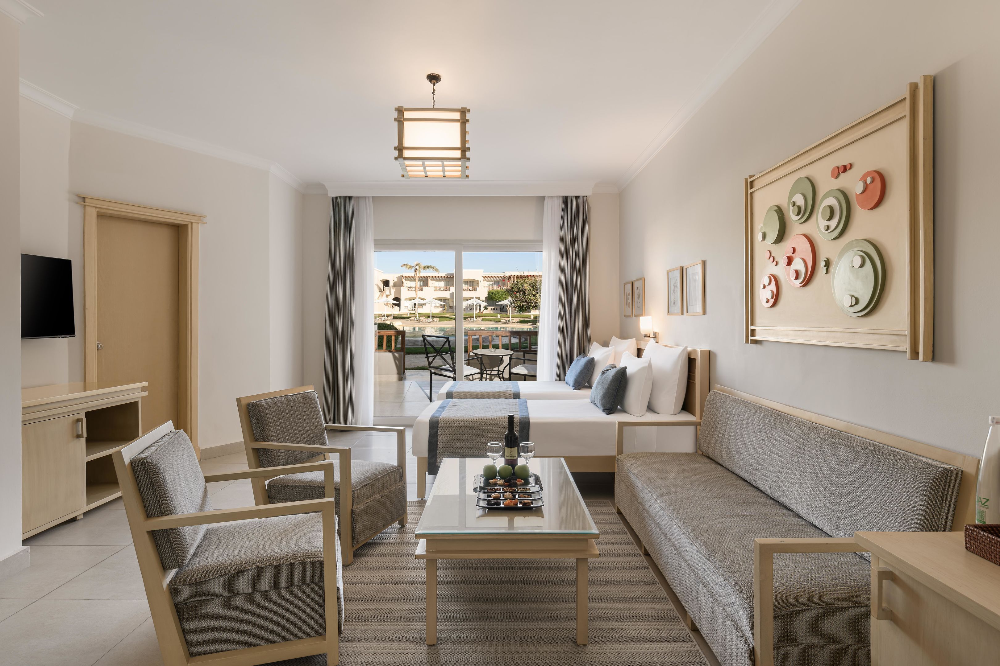 A modern hotel room with a bed, sofa, chairs, a coffee table, and a TV.