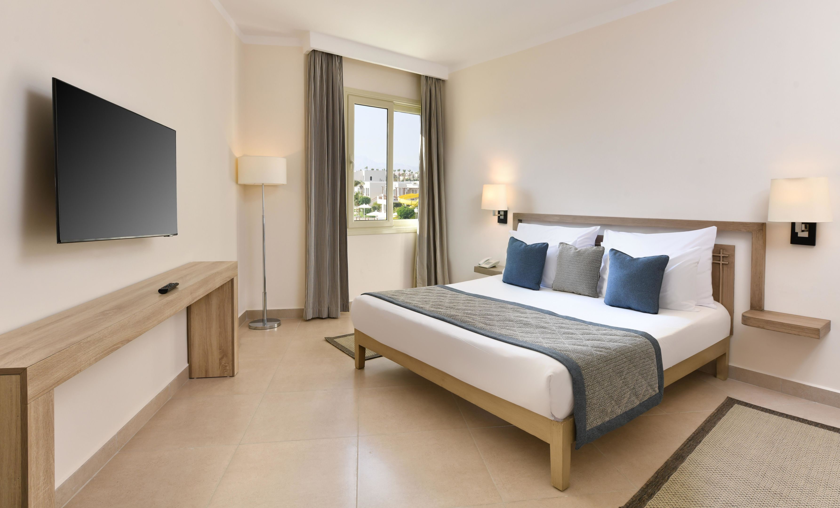 Habitación de hotel con cama, televisión, escritorio, lámpara y vistas al exterior.