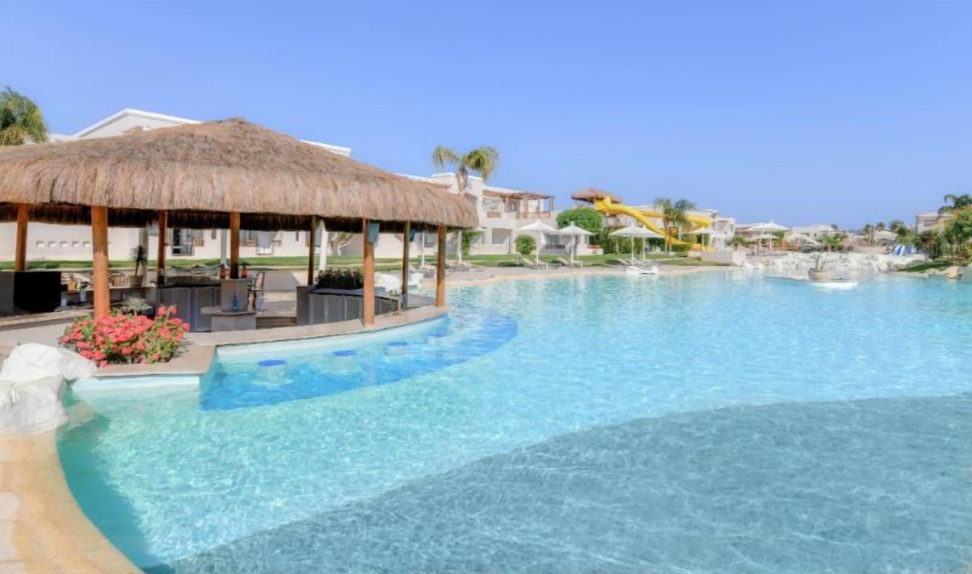 A clear swimming pool with a thatched-roof structure, lounge chairs, and a water slide.