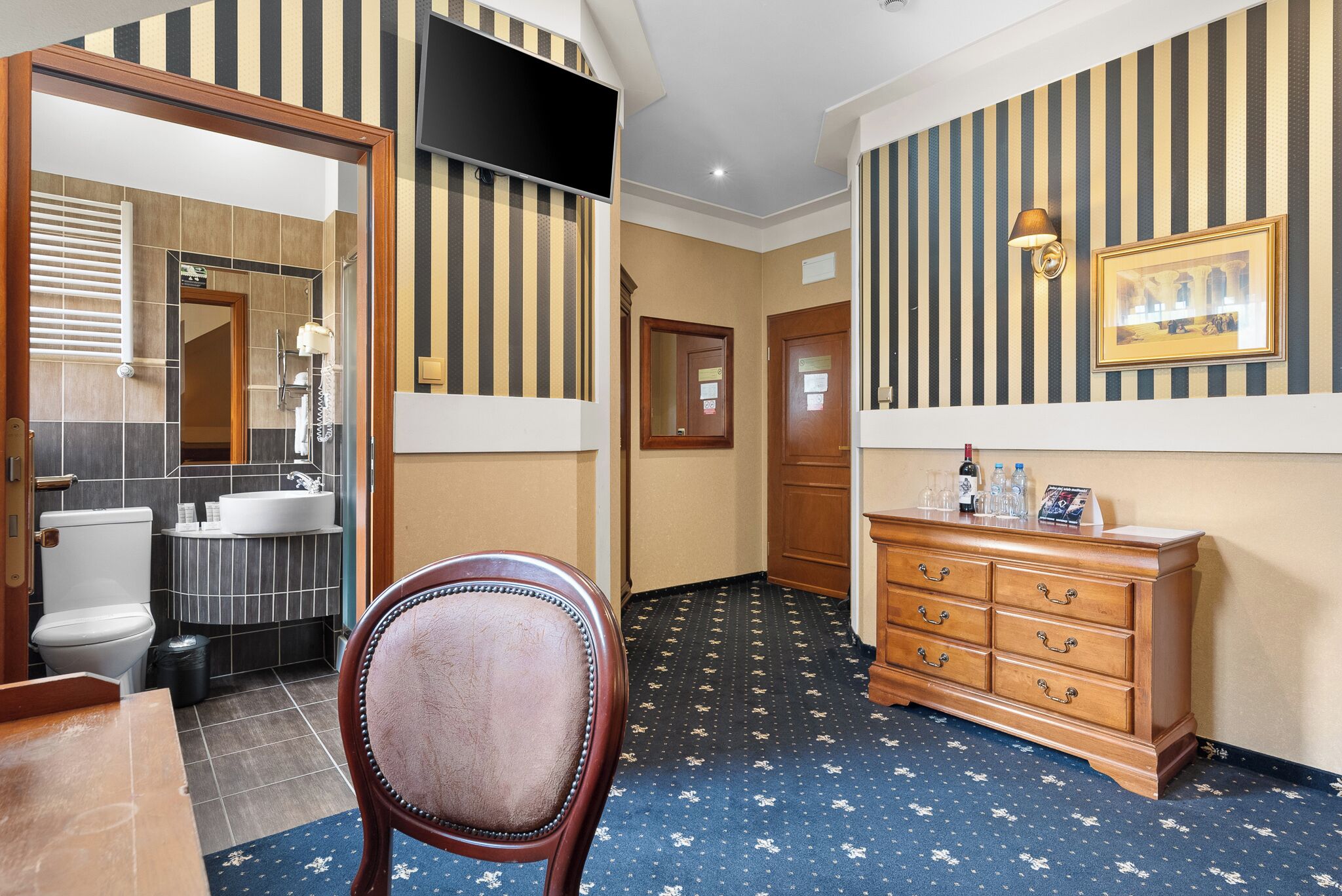 Quarto de hotel com banheiro, penteadeira de madeira, espelho e tapete estampado.