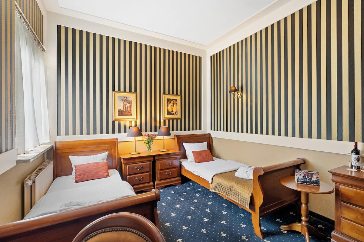A hotel room with two beds, a small table, a nightstand, and a wall-mounted lamp.