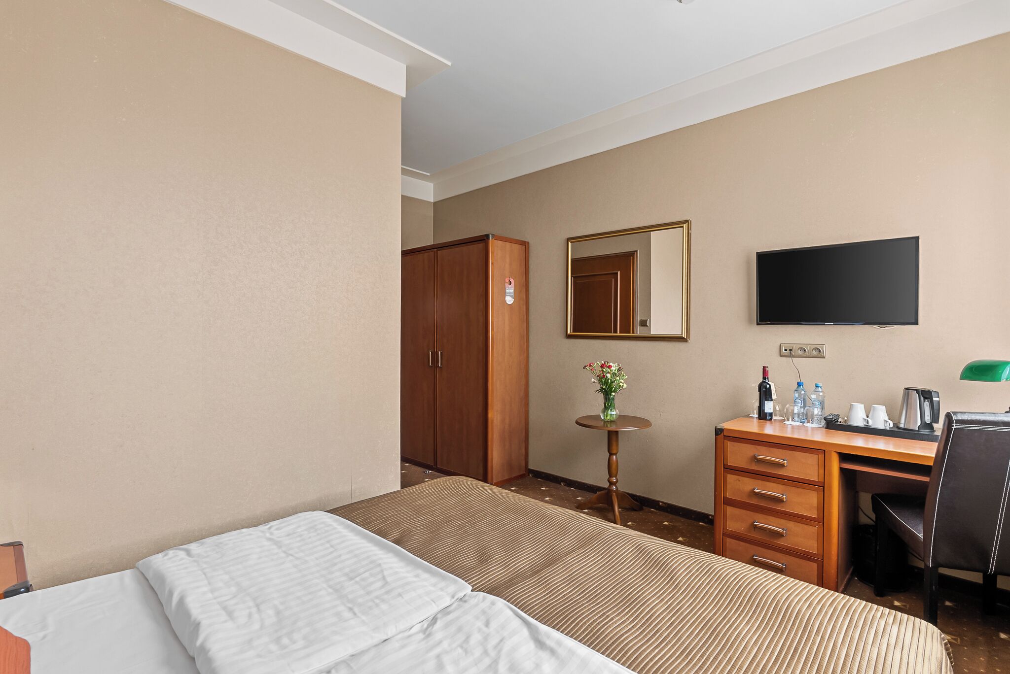 A hotel room with a bed, a desk with a chair, a TV, a wardrobe, and a small table with a vase of flowers.