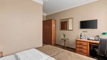 A hotel room with a bed, a desk with a chair, a TV, a wardrobe, and a small table with a vase of flowers.