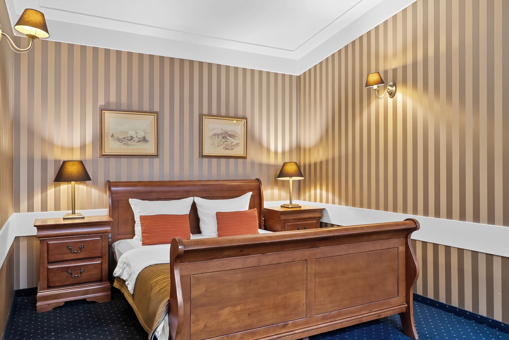A hotel room with a large wooden bed, two bedside tables with lamps, striped wallpaper, and two framed pictures on the wall.