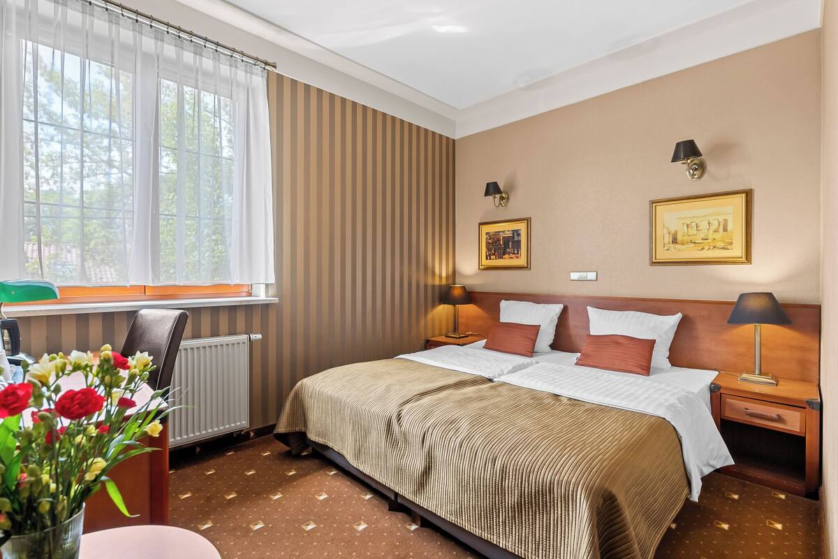 A hotel room with a large bed, two bedside lamps, a window with curtains, a radiator, and a vase with flowers on a table.