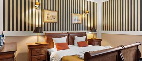 Une chambre d’hôtel avec un grand lit, une tête de lit en bois, deux tables de chevet avec des lampes, une table de nuit et un papier peint à rayures.