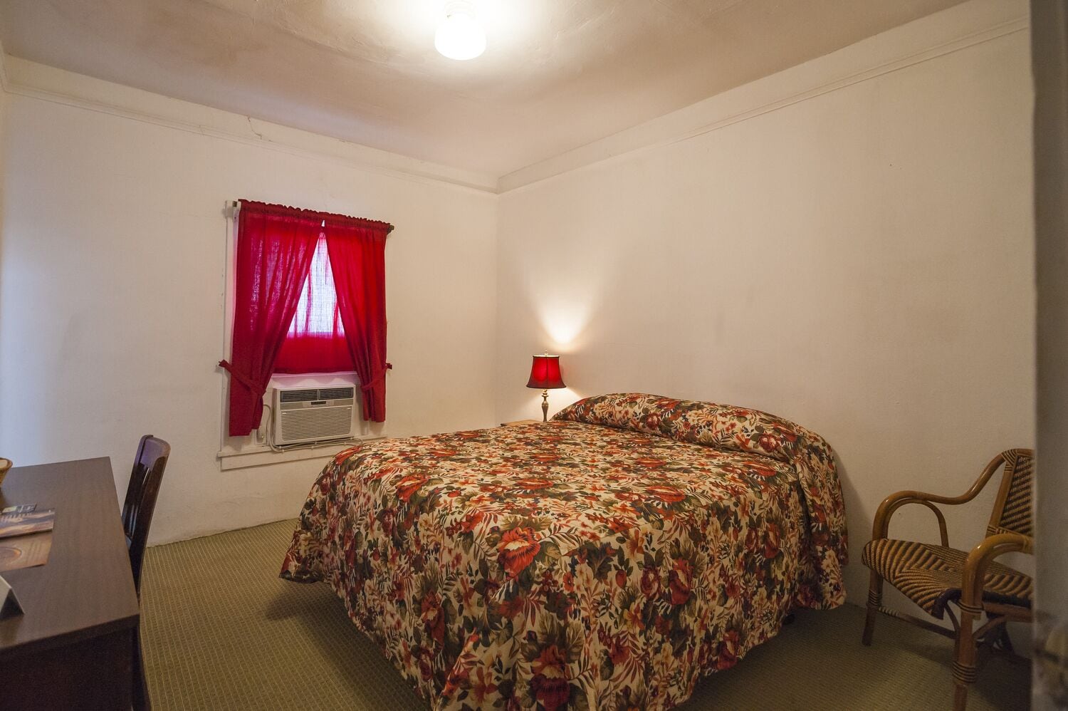 A room with a bed, a desk, a chair, a window with red curtains, and a wall-mounted lamp.