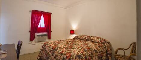 A room with a bed, a desk, a chair, a window with red curtains, and a wall-mounted lamp.