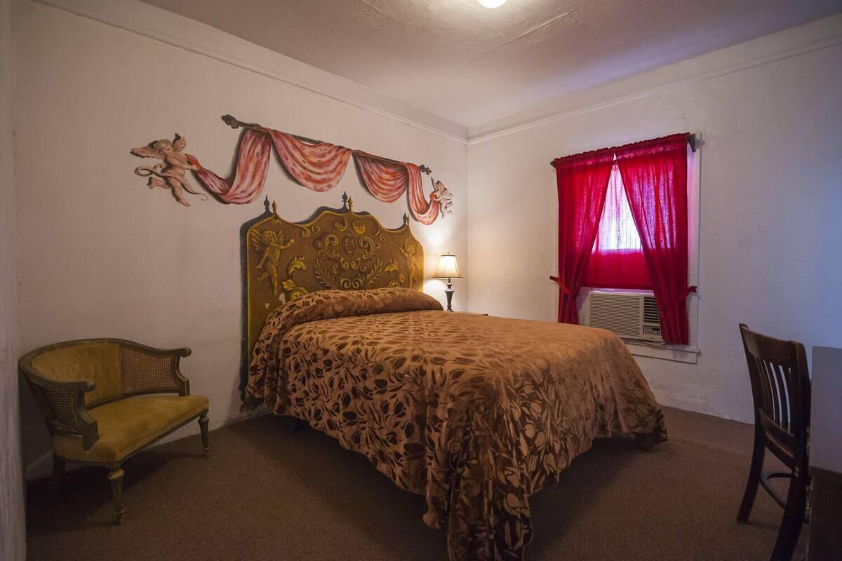 A bedroom with a bed, a chair, a lamp, and a window with red curtains.