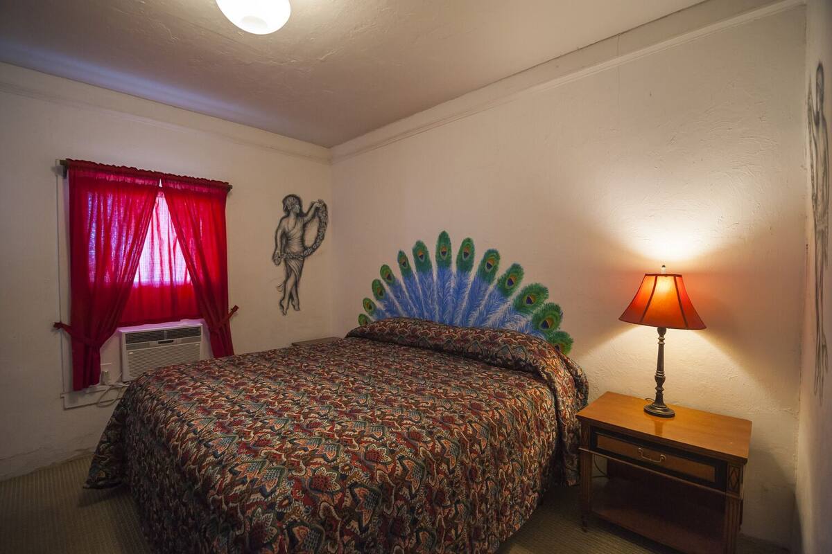 A bedroom with a bed, a peacock feather headboard, a red curtain, a wall clock, and a bedside lamp.