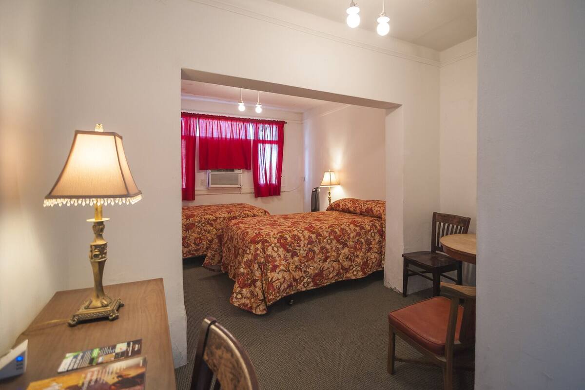 A hotel room with two beds, a desk with a lamp, a chair, and a small table.