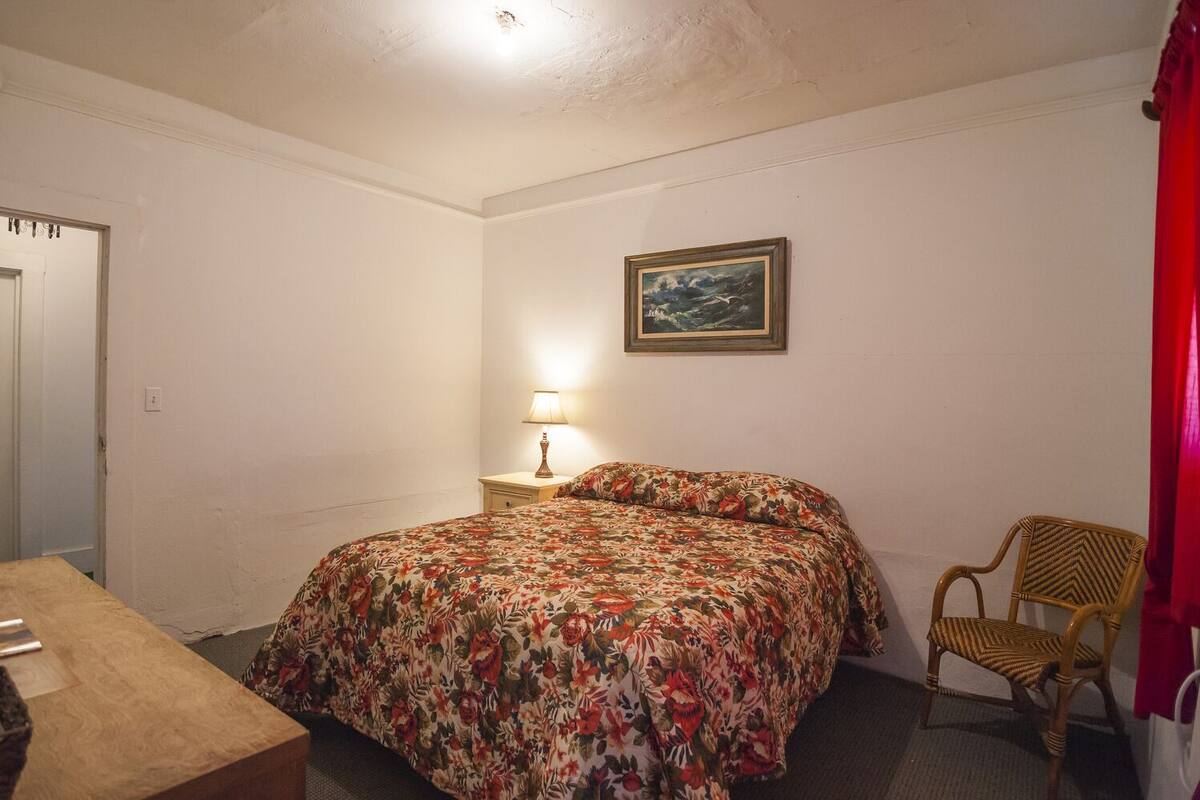 A bedroom with a floral bedspread, a bedside lamp, a chair, and a painting on the wall.