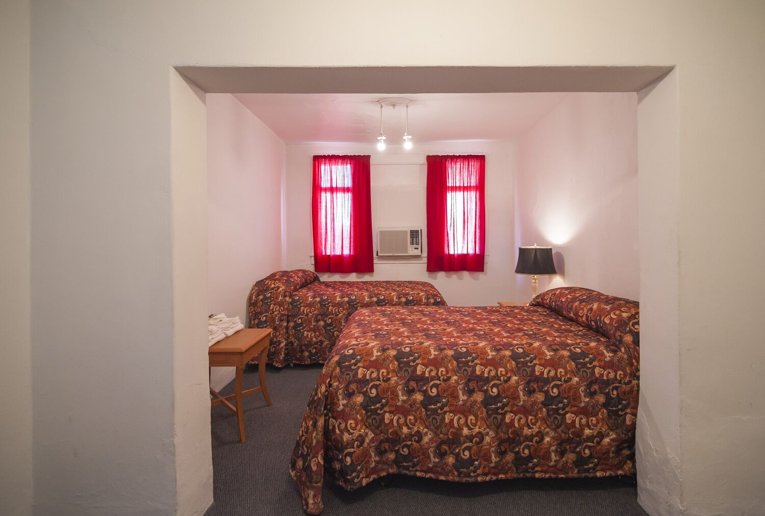 A small, single-room hotel room with two beds, a desk, and a lamp.