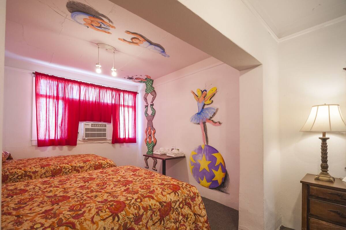 A bedroom with two beds, a window with red curtains, a wall with colorful artwork, and a lamp on a nightstand.