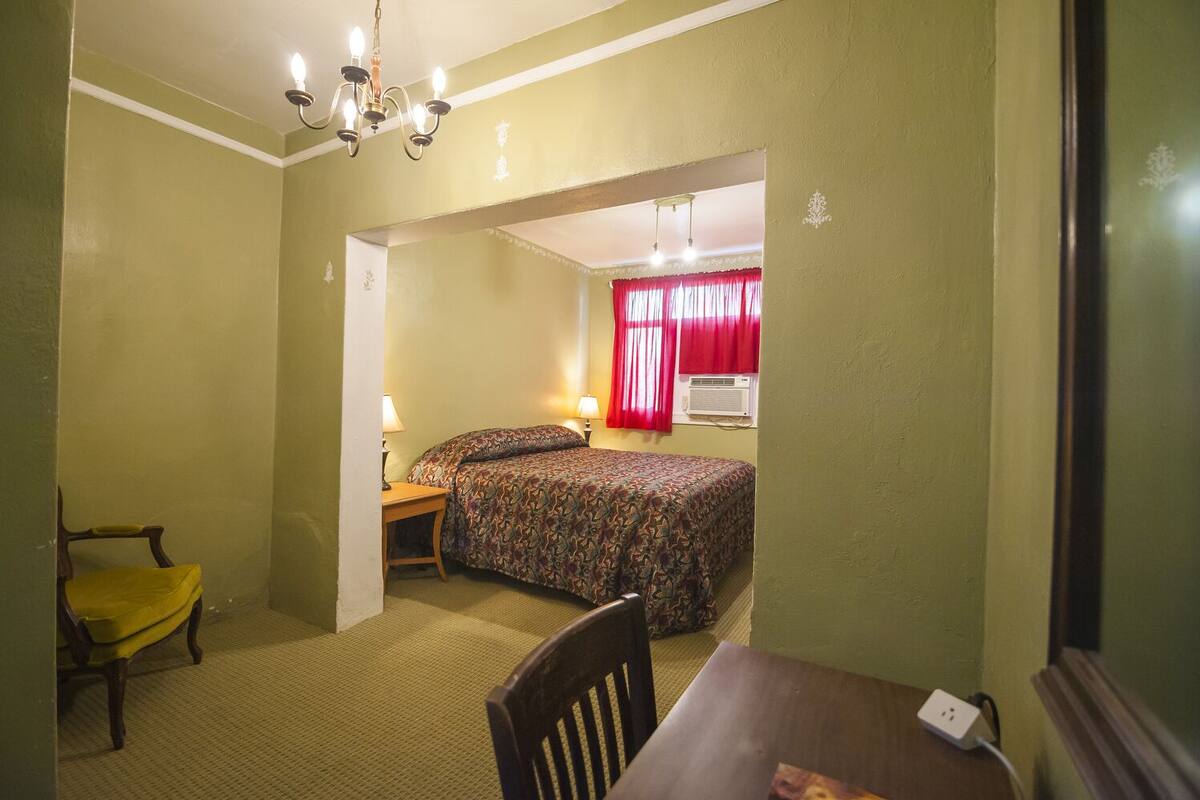 A small, single-room hotel room with a bed, a desk, a chair, and a table. There is a window with red curtains and an air conditioning unit mounted on the wall.