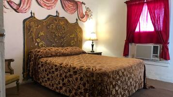 Een keurig opgemaakt bed met een bloemenprintdeken, een klassiek hoofdeinde met decoratieve gravures, een bedlampje en een raam met rode gordijnen.