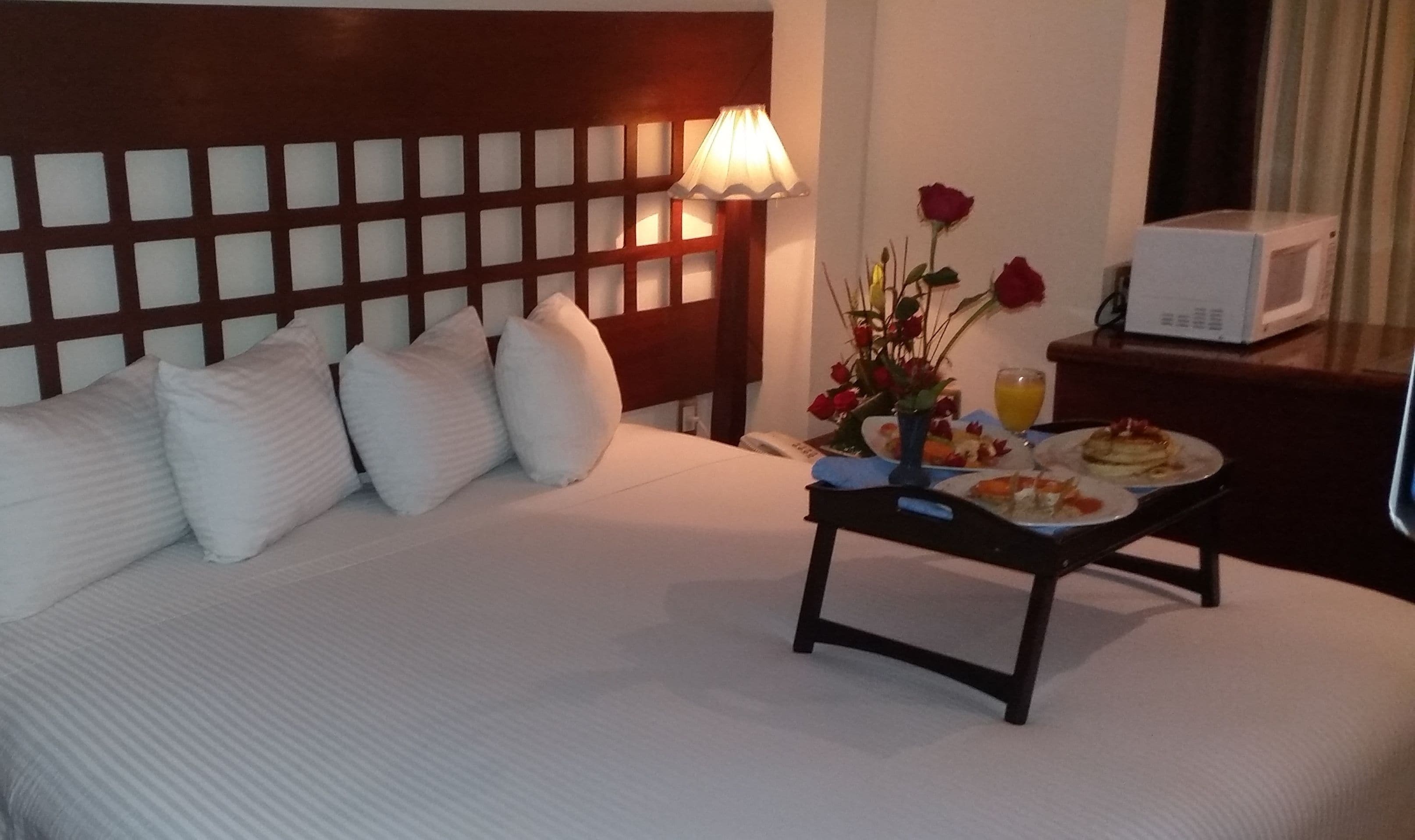 A hotel room with a bed, pillows, a bedside table with a lamp, a vase with flowers, and a tray with breakfast items.