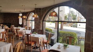 Restaurante com paredes de pedra, amplas janelas em arco e luminárias pendentes. Mesas com toalhas brancas e cadeiras.