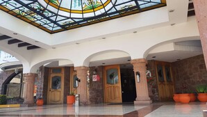 Entrada de hotel com teto de vidro, pilares de pedra e piso de azulejos.