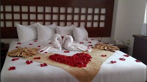 Uma cama bem arrumada com roupa de cama branca, dois travesseiros em formato de cisne e pétalas de rosas vermelhas dispostas sobre o lençol.