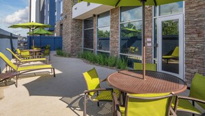 An outdoor patio with yellow chairs and green umbrellas, a round table, and a building with stone and glass windows. - SpringHill Suites by Marriott Lake Charles (Lake Charles)