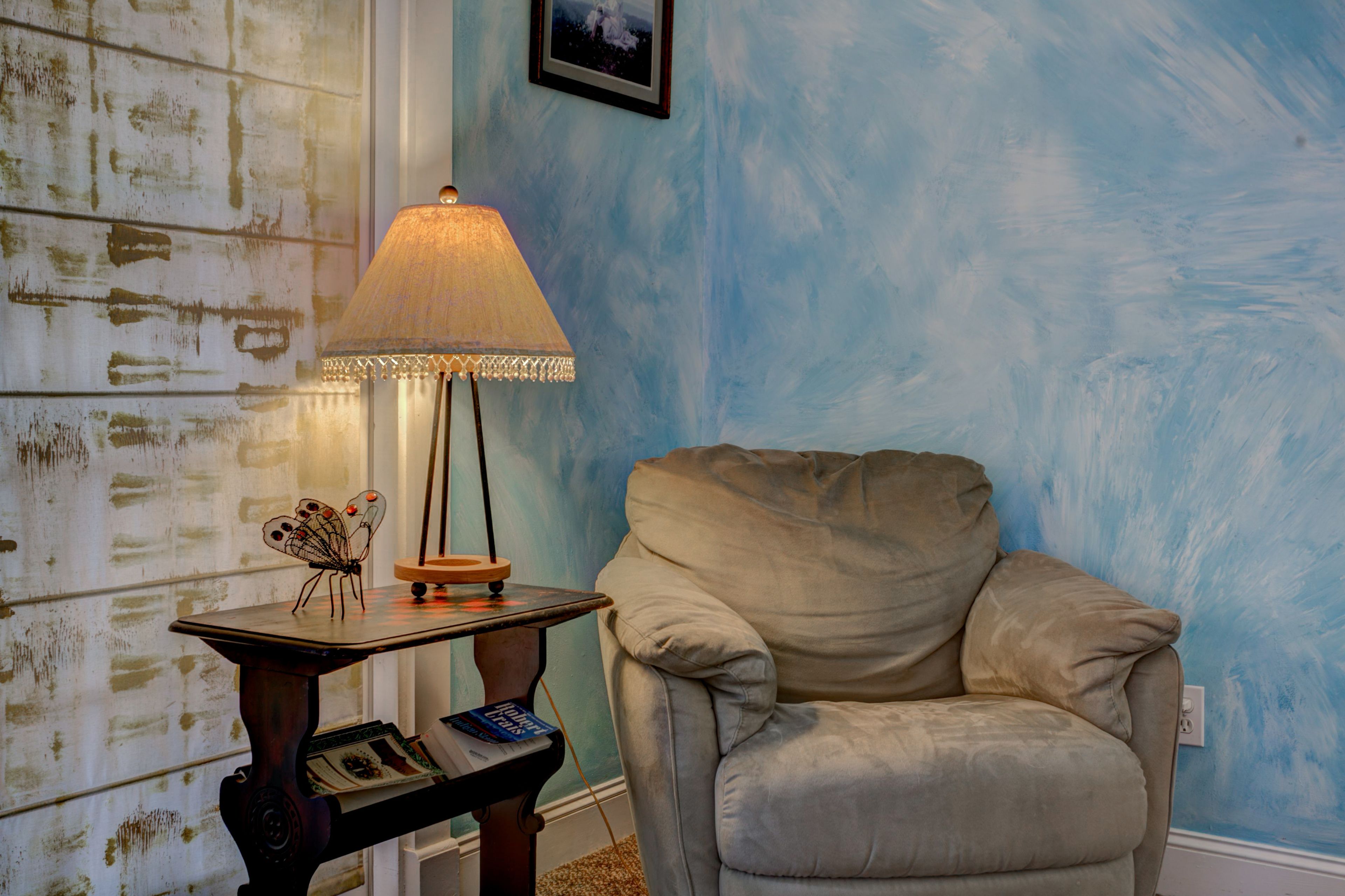 Una habitación con pared azul, un sillón color beige, una mesa con lámpara y un cuadro enmarco.