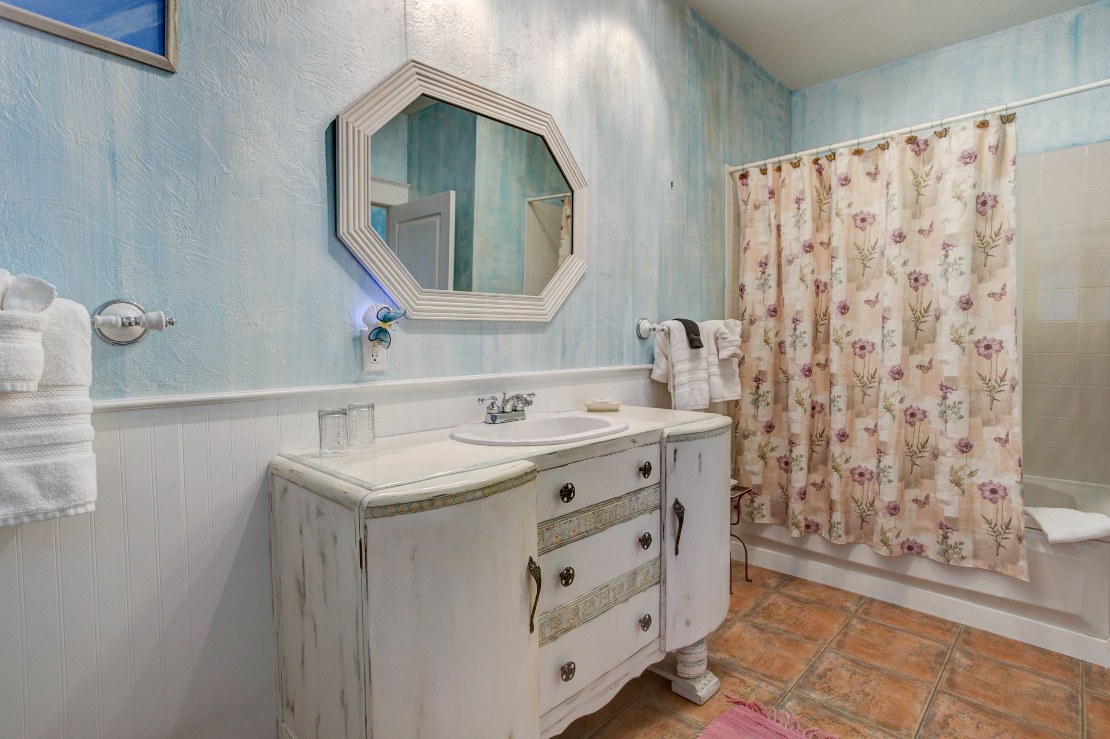 A bathroom with a white vanity, a round mirror, a floral shower curtain, and a tiled floor.