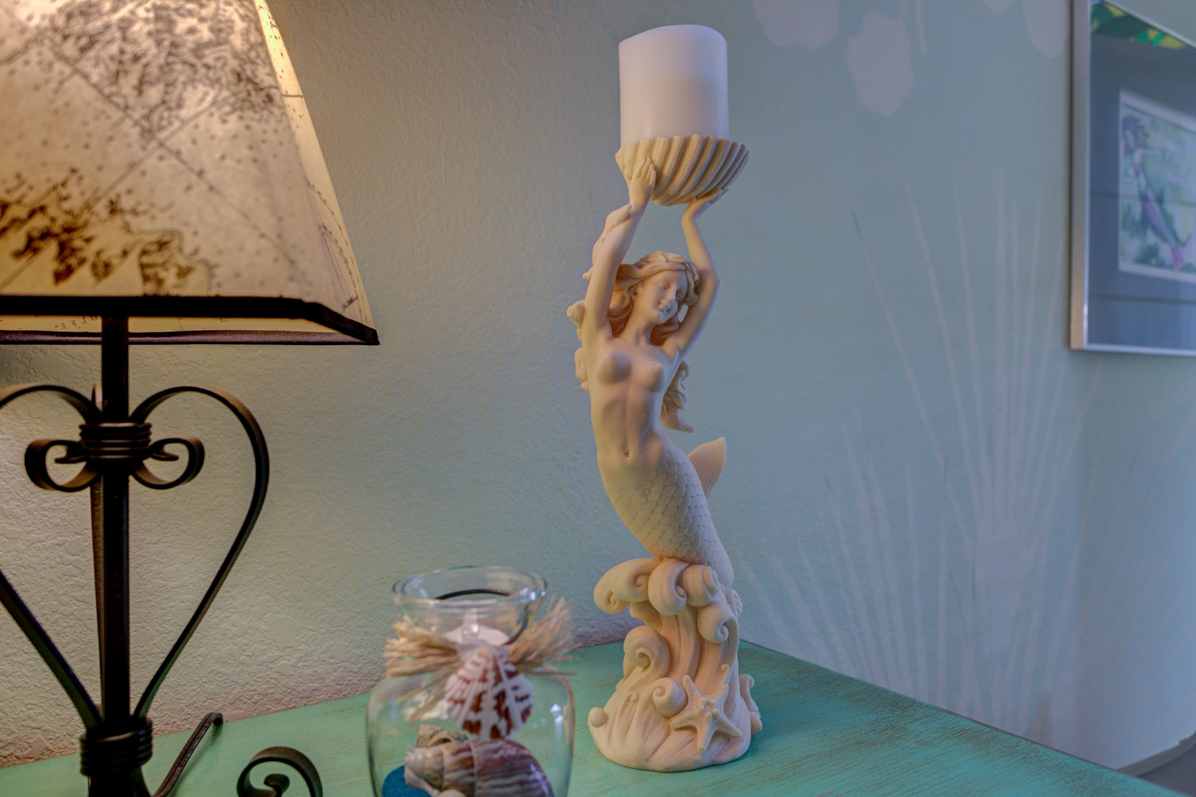 A decorative ceramic mermaid sculpture on a teal table, a glass vase with a seashell, and a lamp with a patterned lampshade.