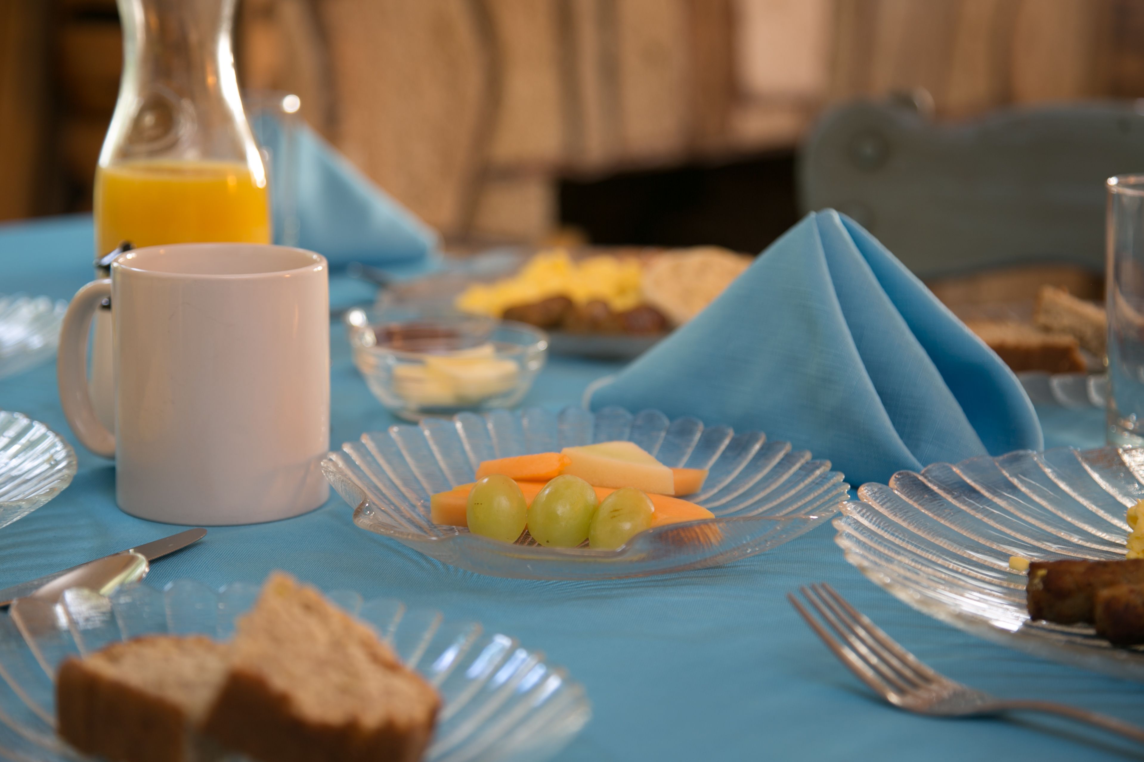 Una mesa de desayuno con una taza blanca, un vaso de jugo de naranja, un plato con queso y uvas, y una servilleta doblada sobre la mesa.