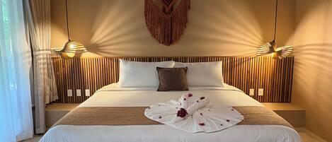 A neatly made bed with a white duvet, a brown throw blanket, and a decorative towel with flowers. There are two wall-mounted lamps on either side of the bed, and a hanging macrame piece above it.