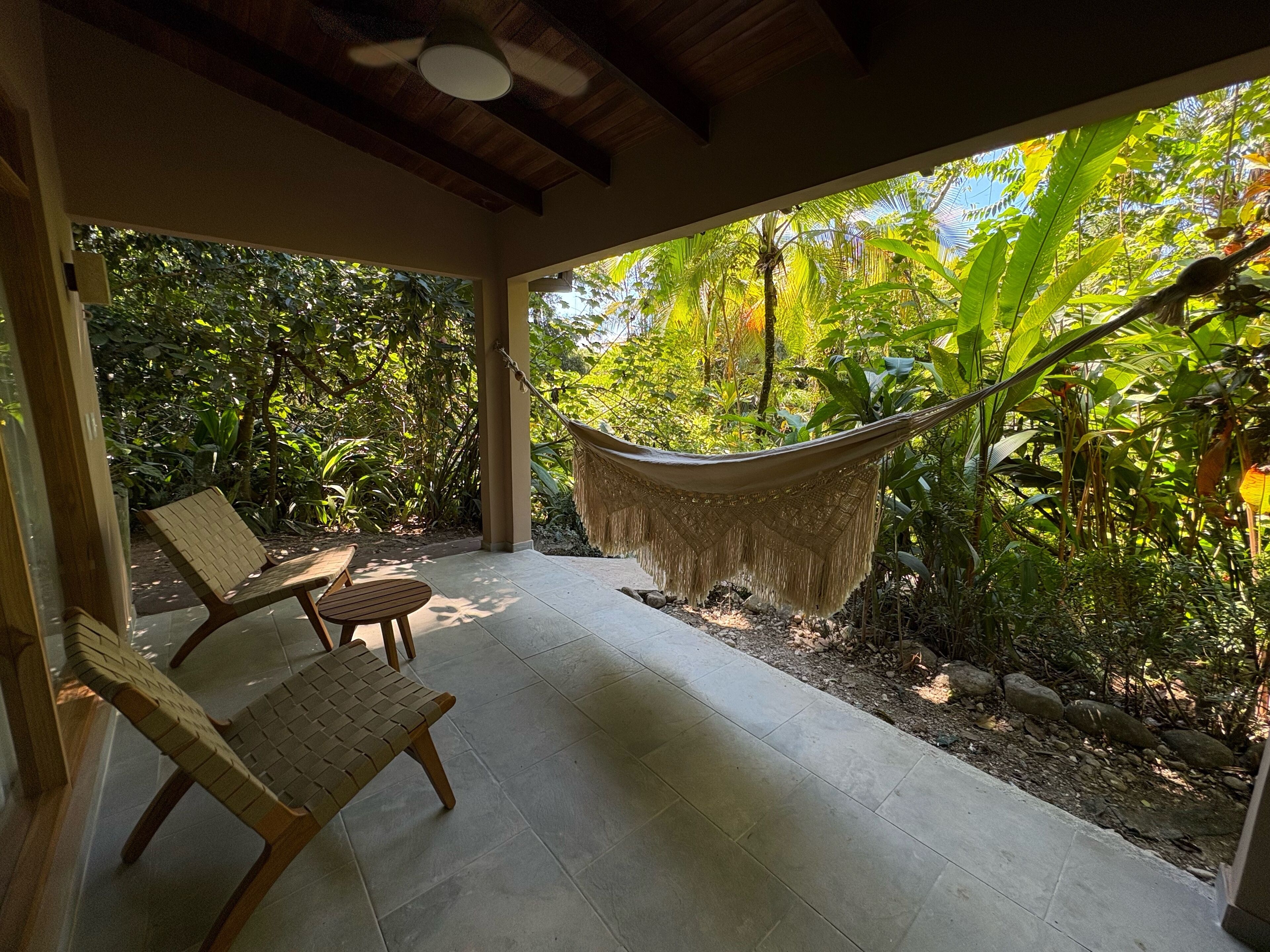 A covered patio with a hammock, two wooden chairs, and a view of lush greenery.