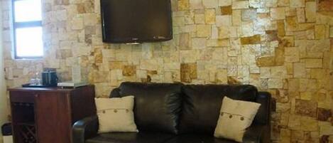 Un salon avec un mur décoré d’un motif imitant la pierre, un téléviseur à écran plat fixé au-dessus d’un canapé en cuir noir agrémenté de deux coussins, et un meuble en bois placé à gauche.