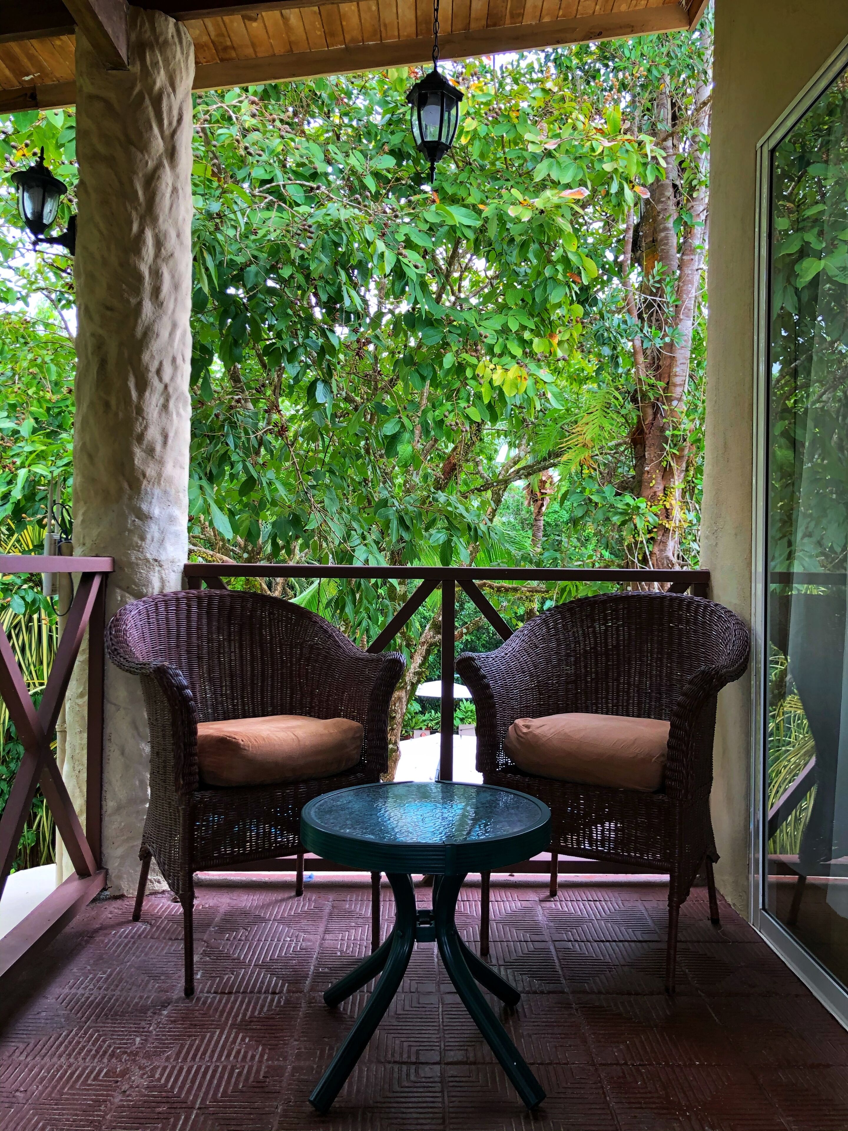 Un espacio de estar techado al aire libre con sillas de mimbre y una mesa de vidrio, rodeado de abundante vegetación.