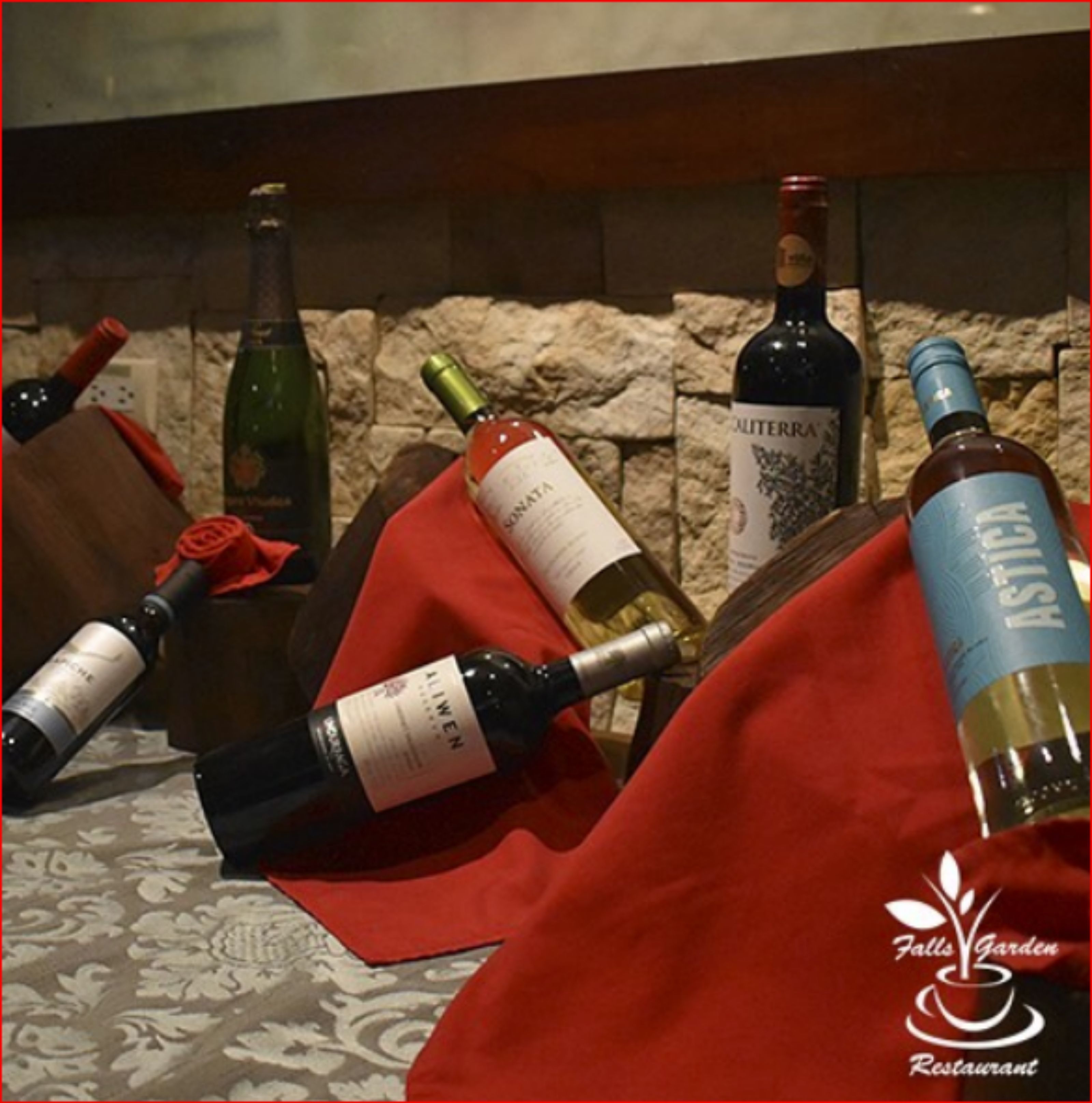 A collection of wine bottles on a table with a red cloth, a stone wall in the background, and a logo for Falls Garden Restaurant.