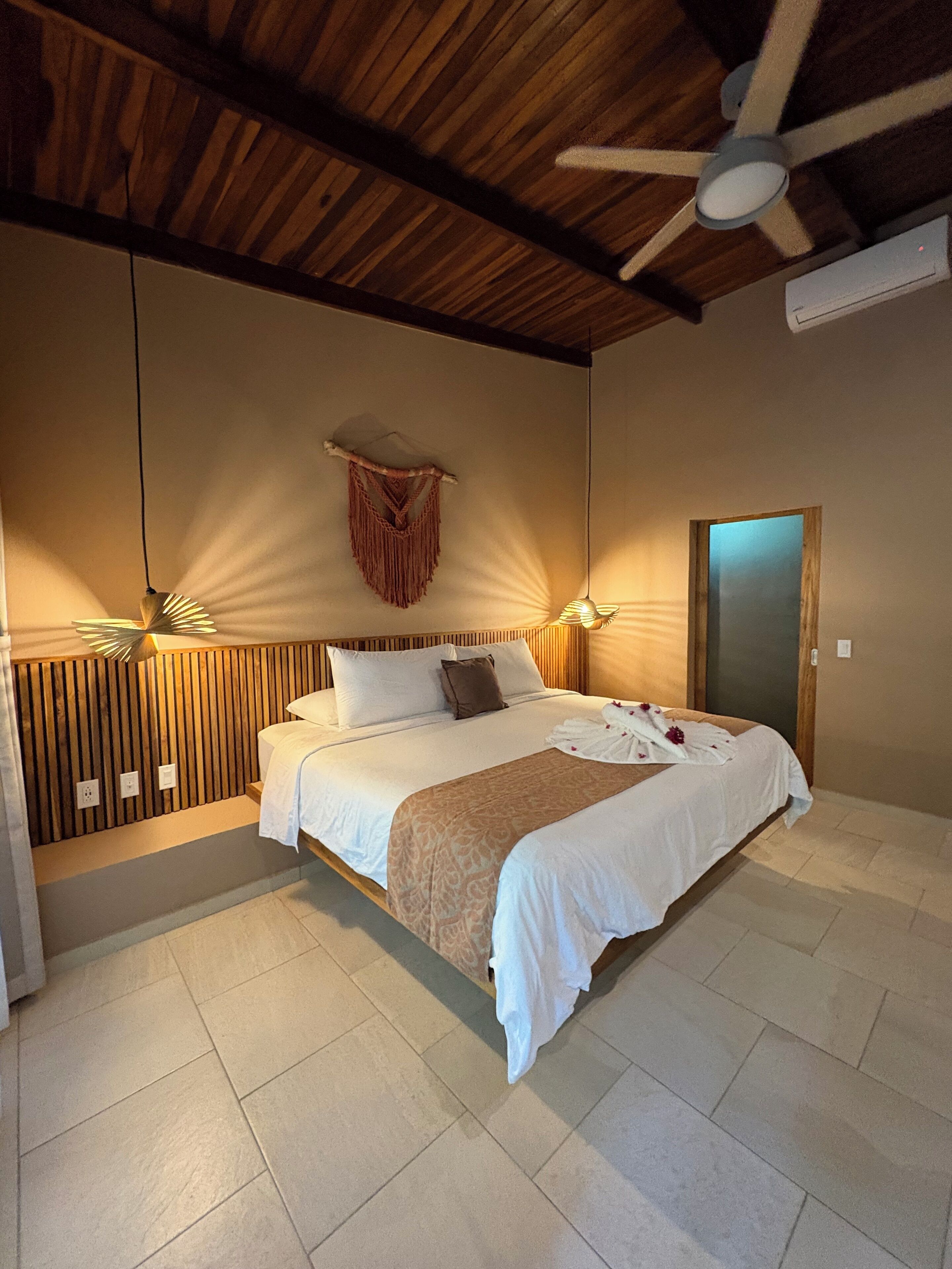 A bedroom with a wooden ceiling, a bed with white bedding, a wooden headboard, a wall-mounted macrame decoration, and a ceiling fan.