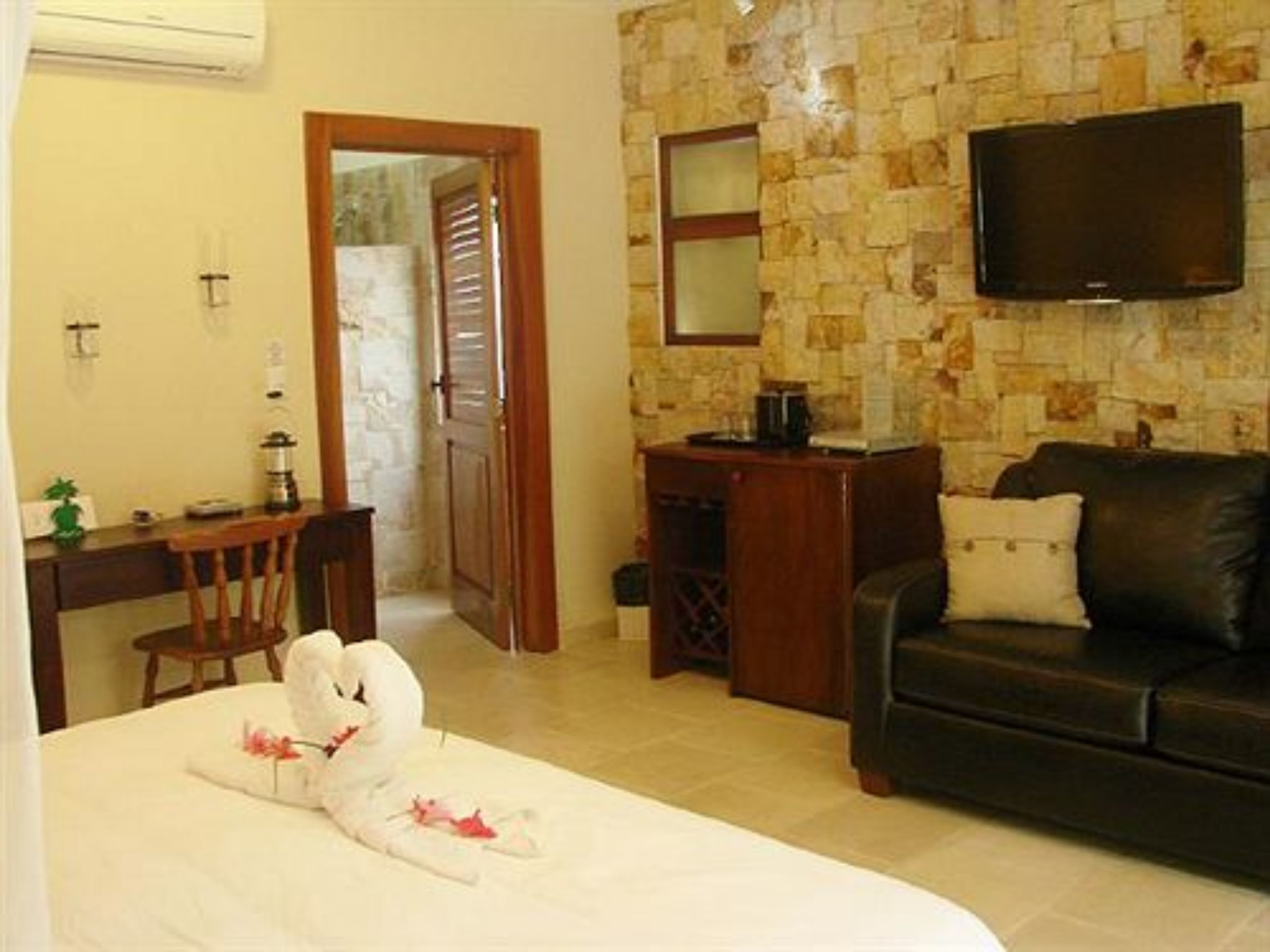 A hotel room with a bed, a wooden desk, a chair, a television mounted on the wall, and a stone-patterned wall.