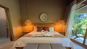 A hotel room with a large bed, two bedside tables, a bench at the foot of the bed, a wall clock, and a window with a view of greenery.