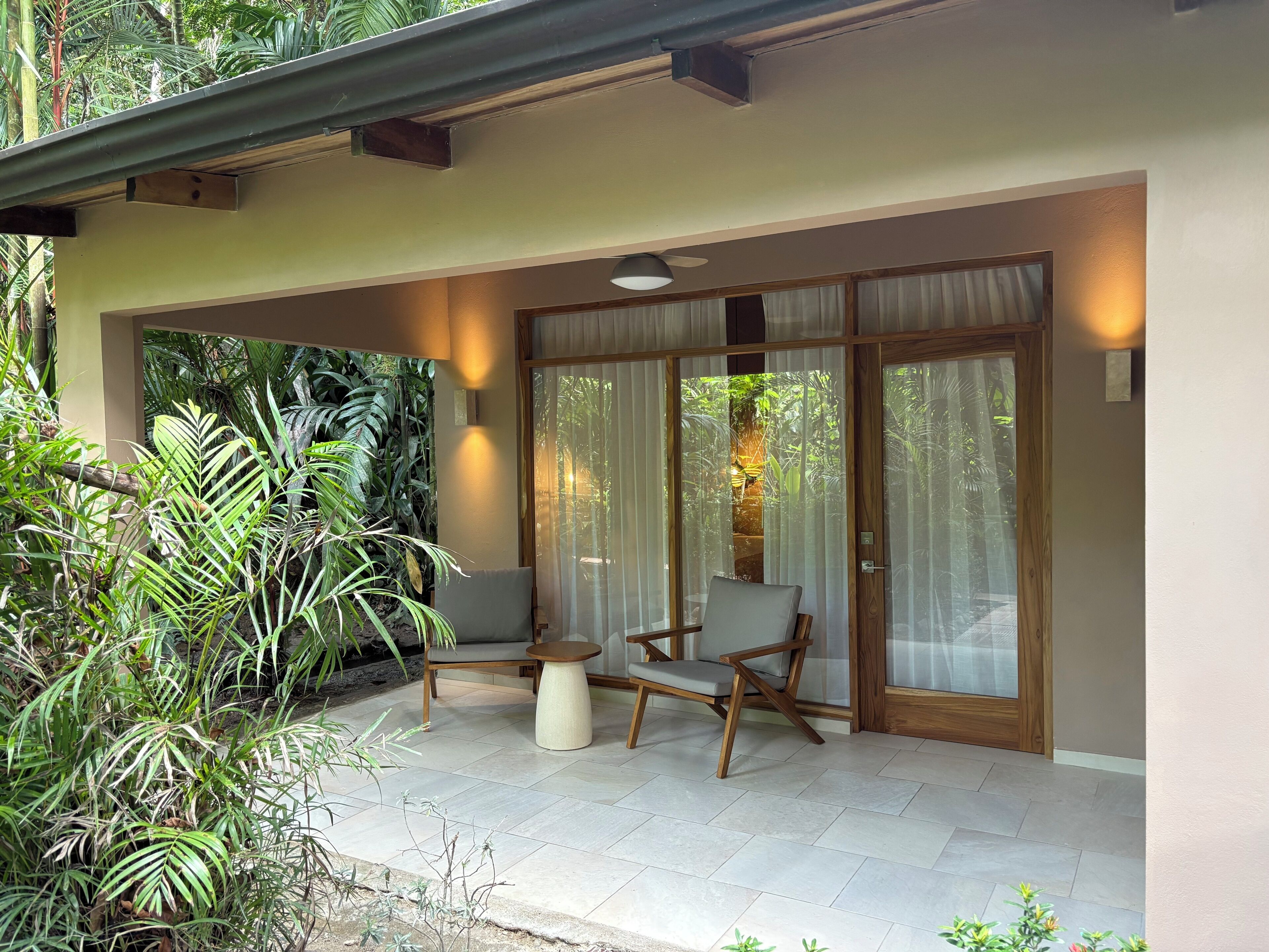 Une terrasse couverte, agrémentée de deux chaises et d’une petite table, entourée de portes et de rideaux en verre, et donnant sur un jardin luxuriant.