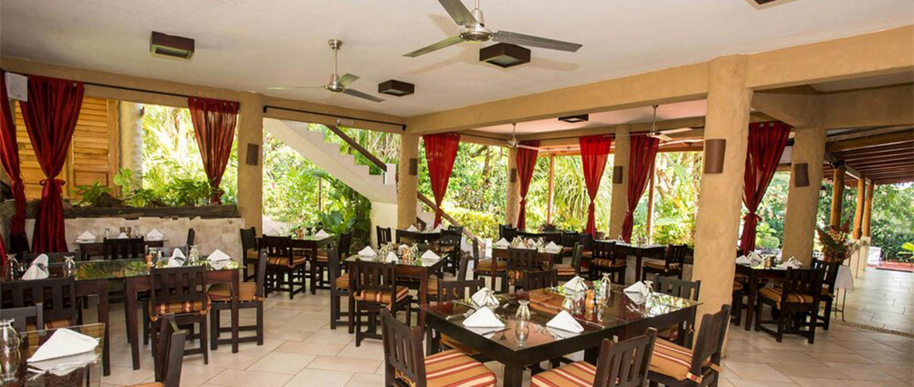 A spacious indoor dining area with multiple tables and chairs, ceiling fans, and large windows with red curtains.