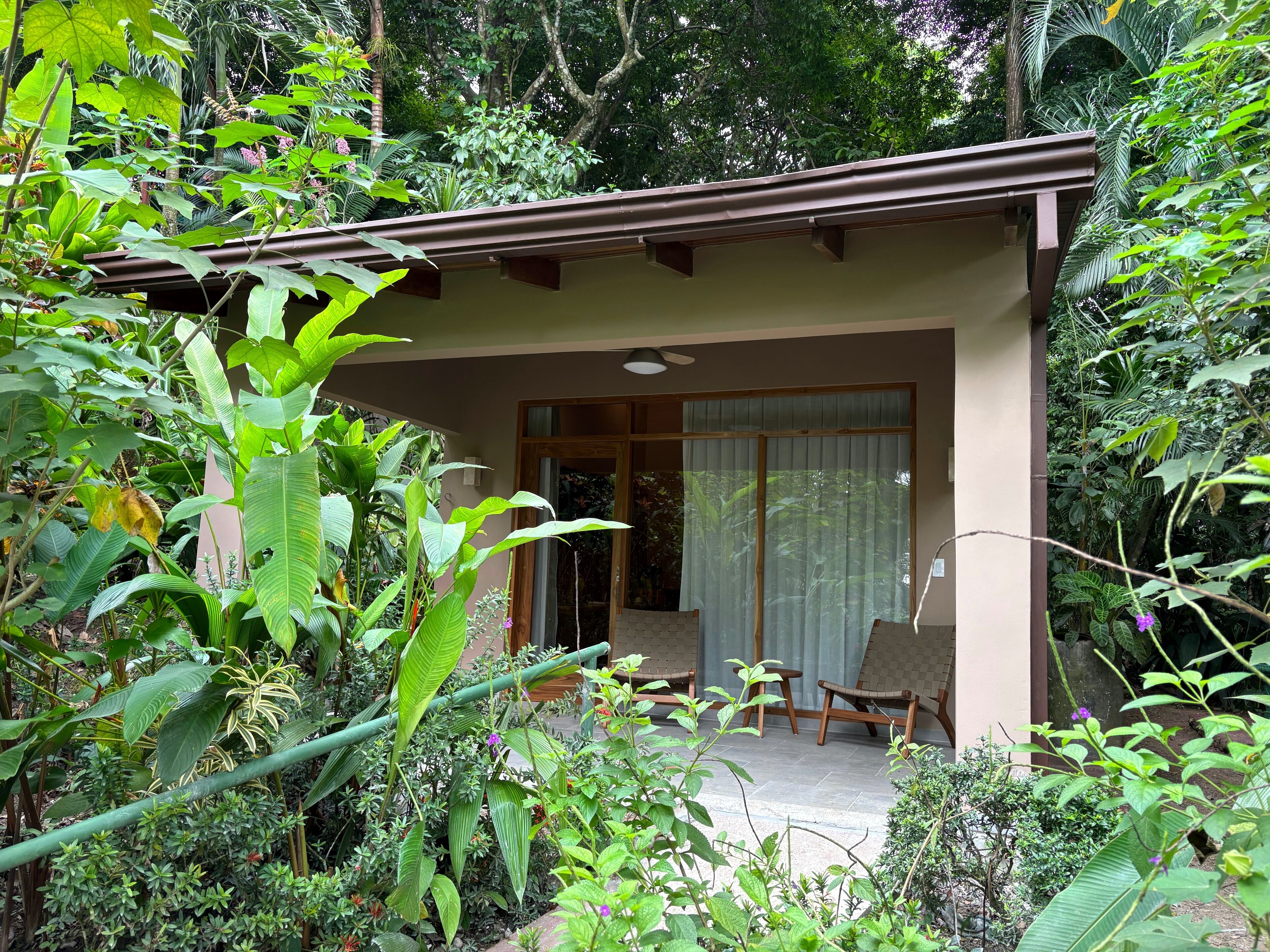 A covered outdoor seating area with chairs and a table, surrounded by lush greenery.